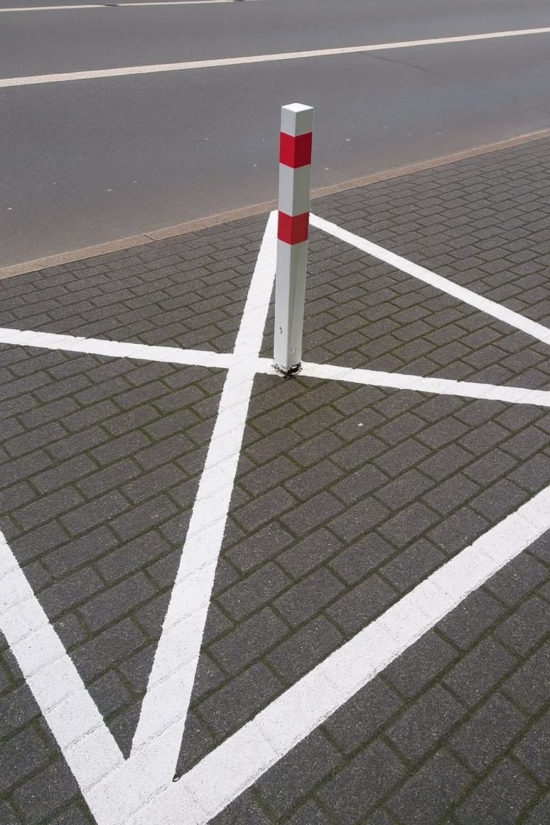 Stĺpik s červenými pruhmi na parkovisku s bielymi čiarami, štandardné dopravné značenie.