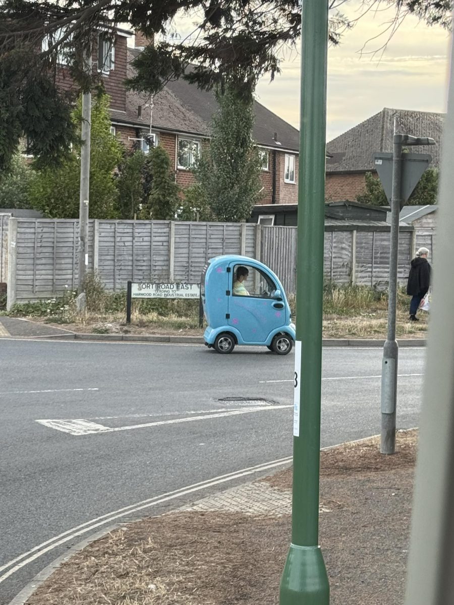 Malé modré vozidlo na ceste v meste, pasažier jazdí popri dome a plote.