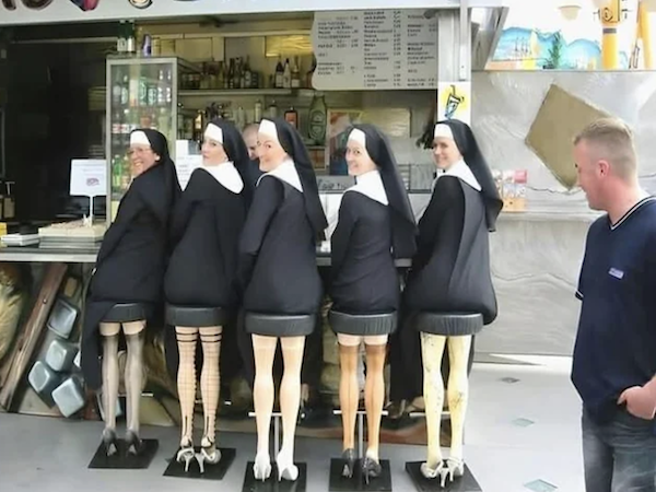 Mníšky s humorom: ženy v kostýmoch pri bare, vyčnievajúca zvláštnosť v podobe nôh na stoličkách.