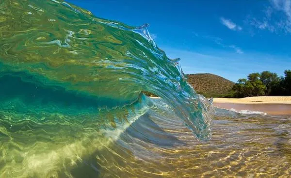 Priezračná vlna naráža na piesočnatú pláž pod modrou oblohou na tropickom pobreží.