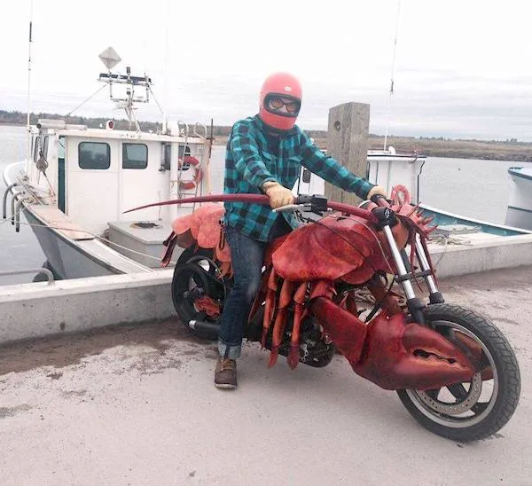 Motorkár na prilbe vedľa motorky v tvare homára, pri móle s rybárskou loďou v pozadí.