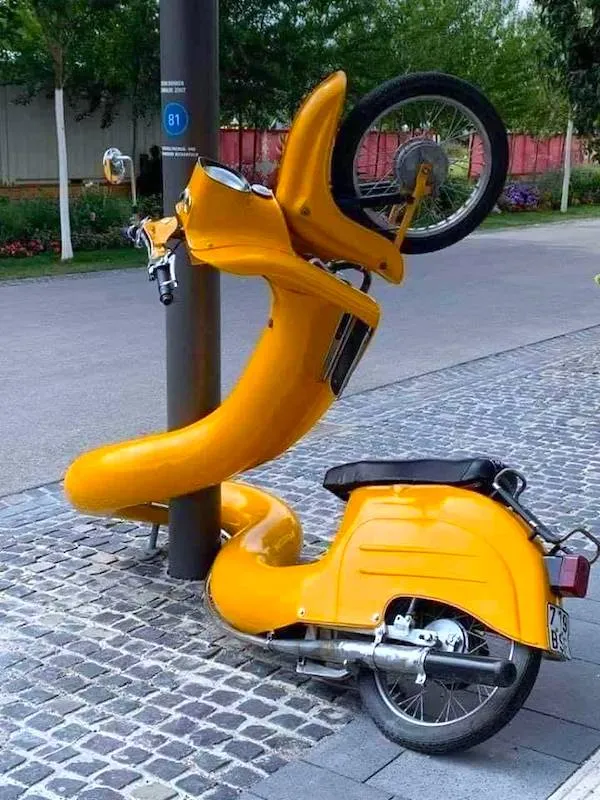 Žltý skúter zauzlený okolo stĺpa na ulici, výrazný umelecký kúsok.