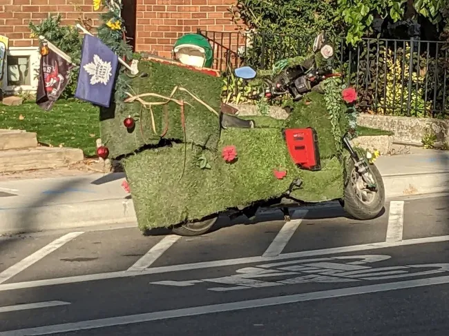 Motorka pokrytá umelou trávou so zdobením, zaparkovaná na ceste pri chodníku, vedľa tehlového múru.