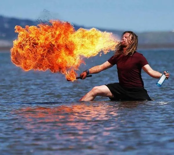 Osoba predvádza ohňovú šou na pláži, sedí vo vode a vypúšťa plamene pomocou aerosólu.