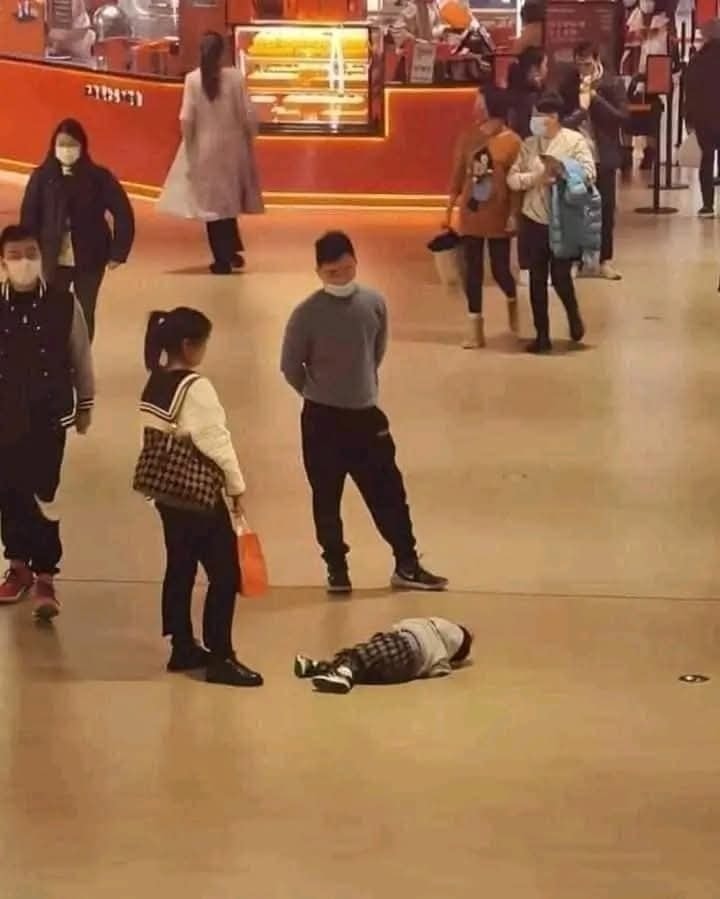 Dieťa leží na zemi v nákupnom centre, okolo neho stoja dospelí a ďalší ľudia v pozadí.