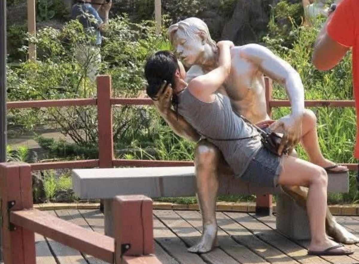 Osoba na lavičke objíma sochu v parku v letnom oblečení, v pozadí zeleň.