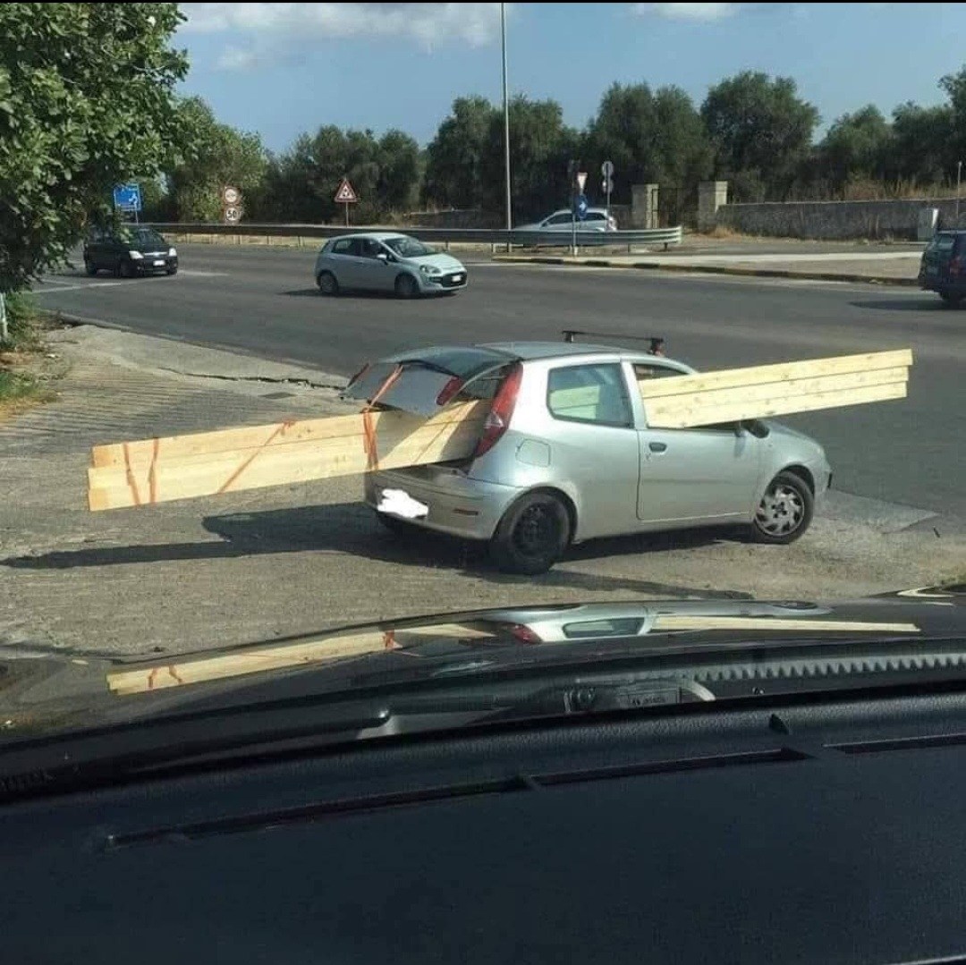 Malé auto prepravuje dlhé drevené dosky, ktoré trčia z oboch koncov vozidla na okraji cesty.