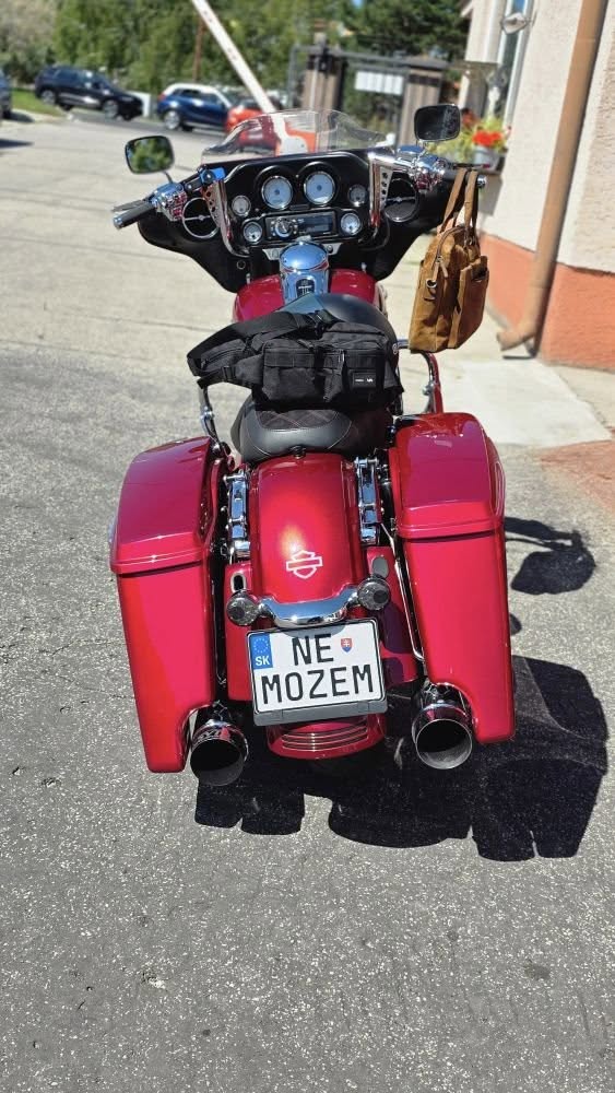 Červená motorka Harley Davidson s tabuľkou NE MOŽEM zaparkovaná pri dome na slnečnej ceste.