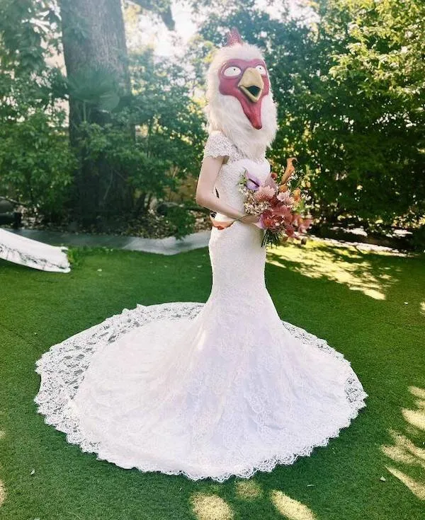 Nevesta v bielej svadobnej šaty s maskou sliepky drží kyticu na zelenej trávnikovej ploche.