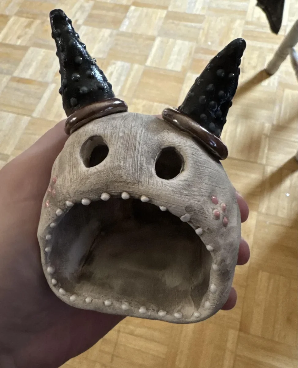 Keramická soška tvora s otvorem úst a čiernymi rohmi držiaca v ruke nad drevenou podlahou.