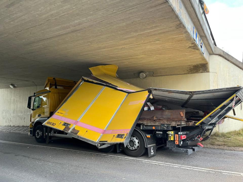 Žltý kamión uviaznutý pod nízkym mostom, poškodená strecha nákladného vozidla.