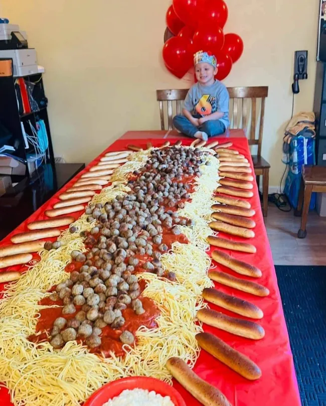 Dieťa sediace pri stole plnom špagiet s mäsovými guľkami a bagetami, dekorované červenými balónmi.