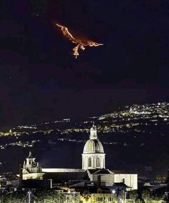 Nočná erupcia sopky nad mestom s osvetlenou katedrálou v popredí, dramatický kontrast svetla a tmy.