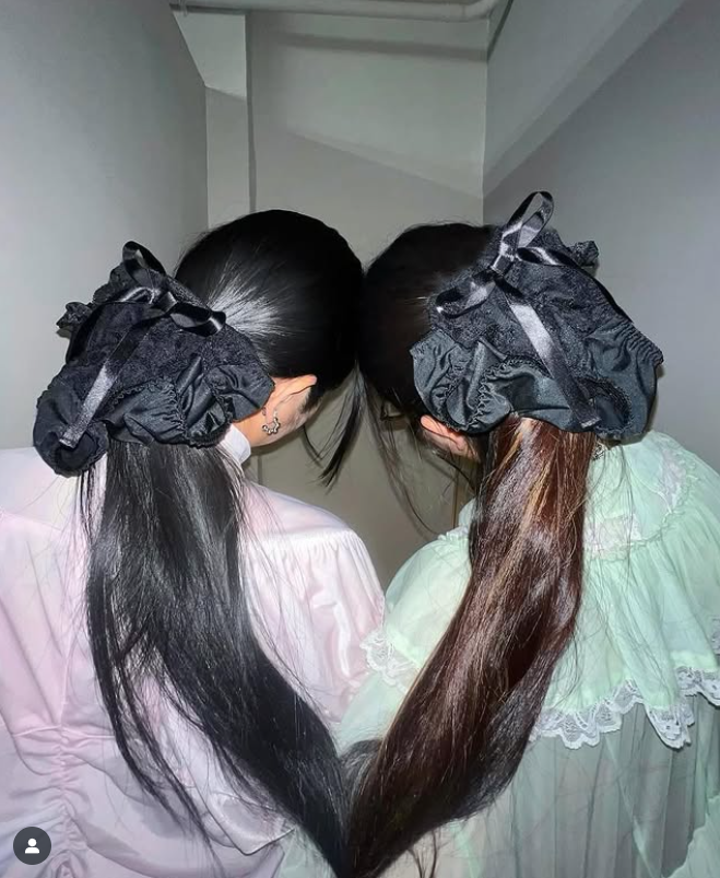 Dve ženy s dlhými vlasmi upravenými do mašlí z čiernej látky, stojace v úzkej chodbe.