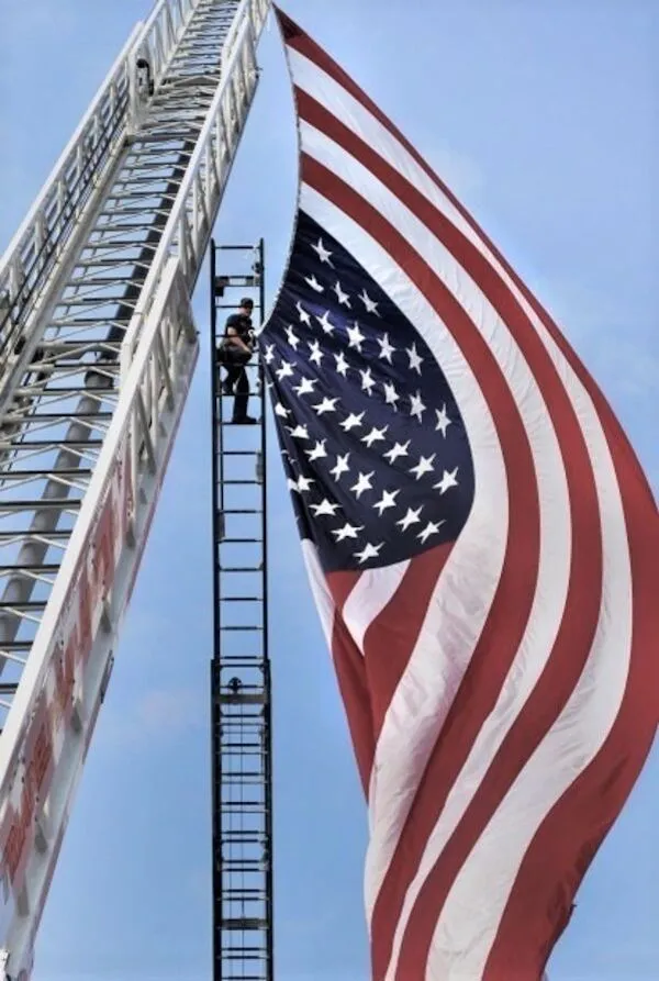 Požiarnik na rebríku vedľa veľkej americkej vlajky, pripravený na zavesenie v modrej oblohe.
