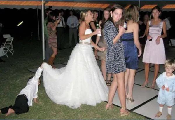 Žena v svadobných šatách tancuje s hosťami, zatiaľ čo dieťa drží jej vlečku na vonkajšej oslave.