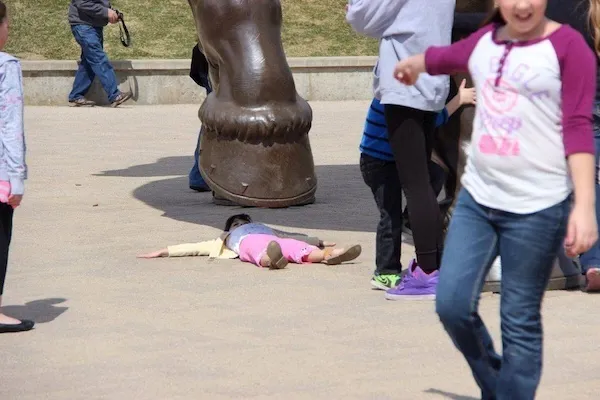 Dieťa leží na zemi pod veľkou sochou v parku, okolo prechádzajú ľudia v bežných oblečeniach.