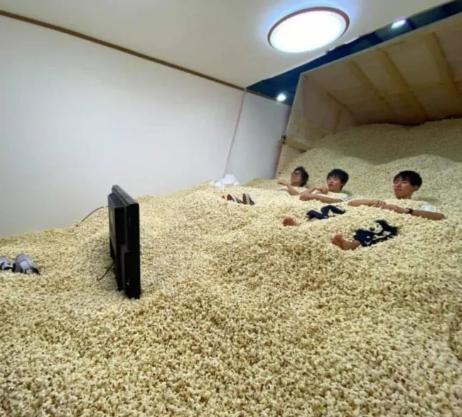 Ľudia sledujú televíziu ponorení do obrovskej hromady pukancov v miestnosti.