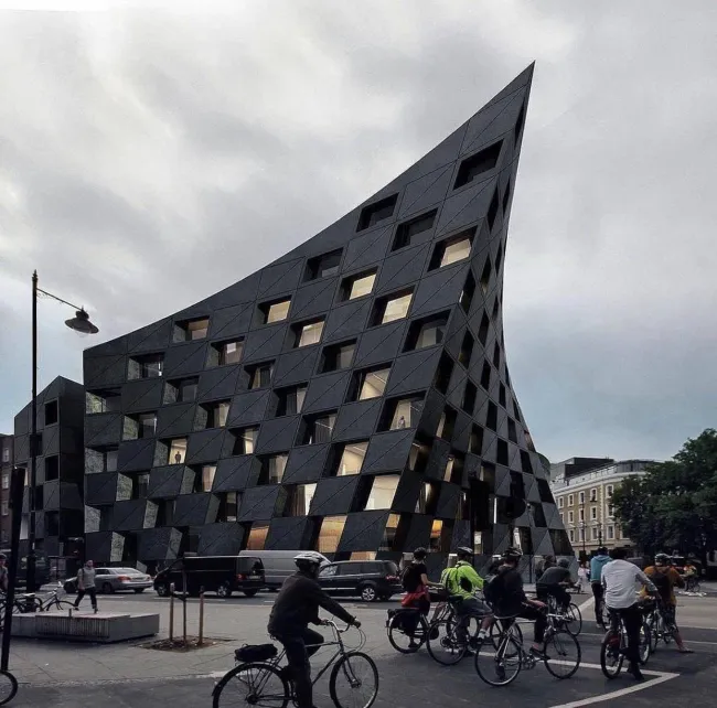 Moderná budova s unikátnou architektúrou, ľudia na bicykloch v rušnom mestskom prostredí.