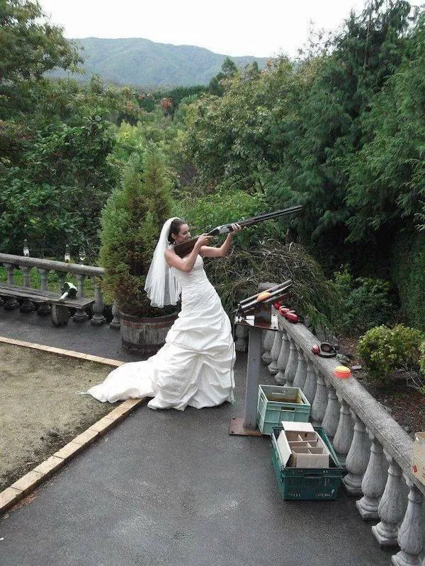 Nevesta v bielych svadobných šatách mieri puškou na terči v prírode, ukazujúc netradičný svadobný moment.