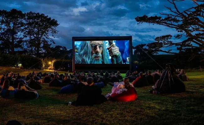 Vonkajšie premietanie filmu na trávniku s ľuďmi sediacimi na dekách a babatách počas západu slnka.