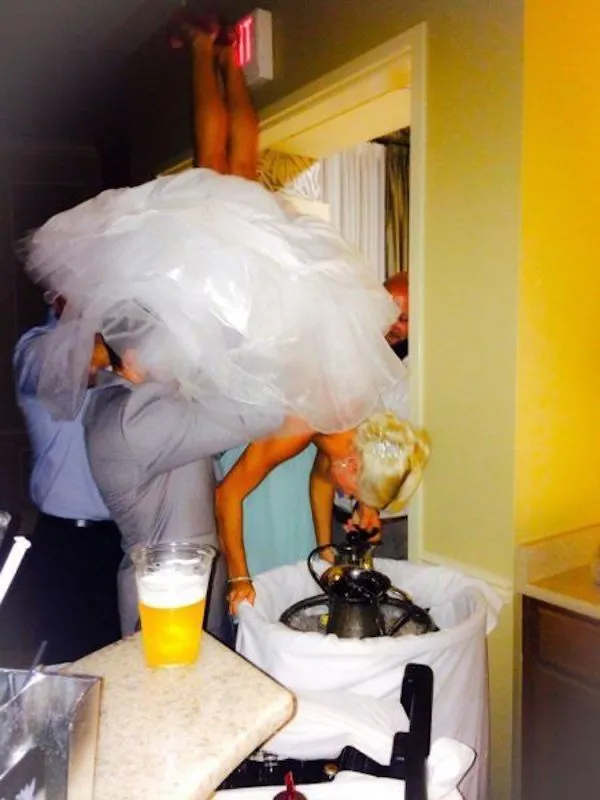 Nevesta v bielej sukni naruby hľadajúca niečo v kôši, obklopená zábavnou atmosférou svadobnej oslavy.