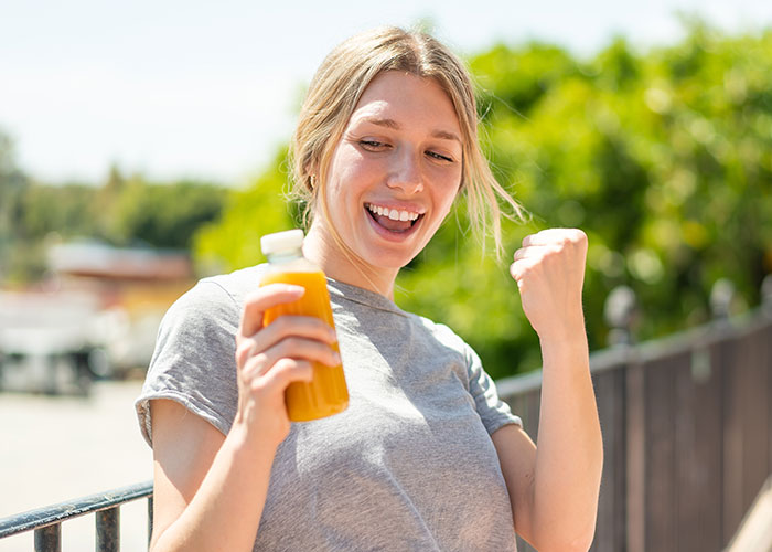 Osoba s úsmevom drží fľašu pomarančového džúsu na slnečnom dni vonku.