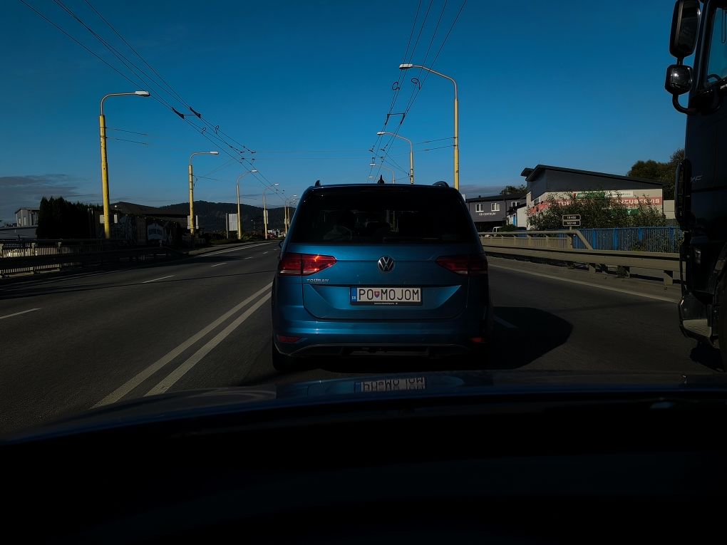 Auto na ceste so špecifickou poznávacou značkou, obloha je jasná a okolo sú mestské budovy.