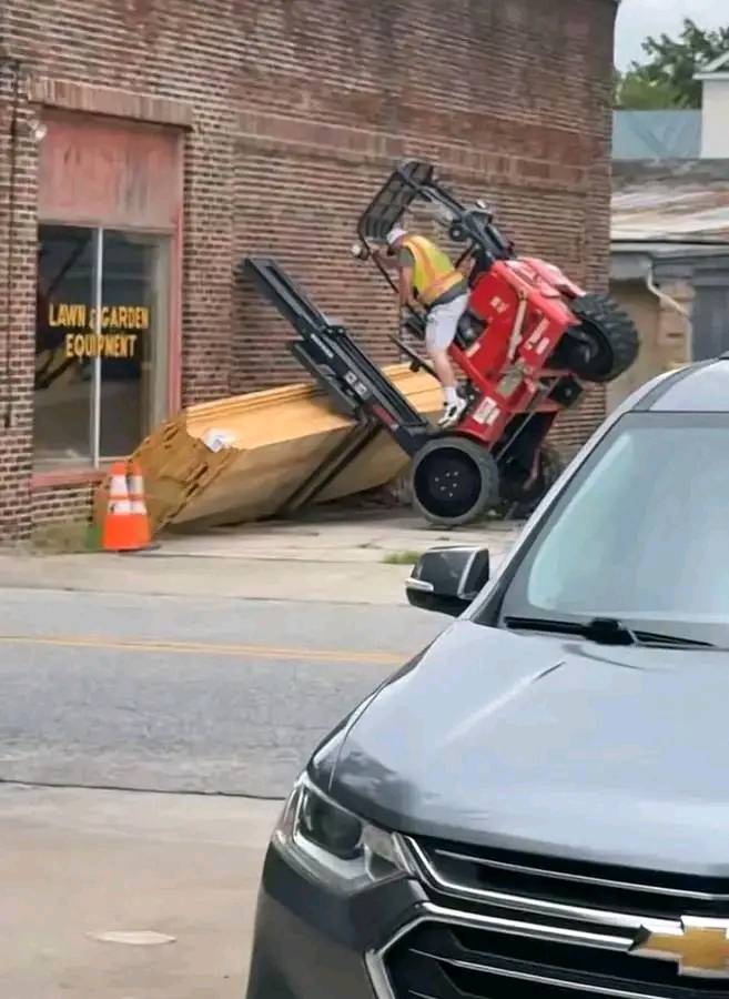 Vysokozdvižný vozík prevrátený proti stene, vodič v reflexnej veste vyzerá zmätene, auto v popredí.