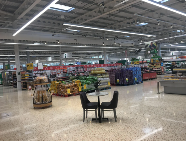 Moderný supermarket s regálmi plnými potravín, stoličky pri stole v popredí, široký priestor na nakupovanie.