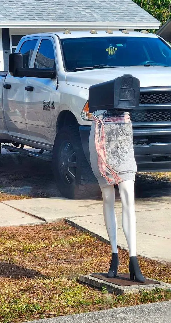 Poštová schránka na figuríne s nohami v šatách pred bielym pick-upom na ceste.