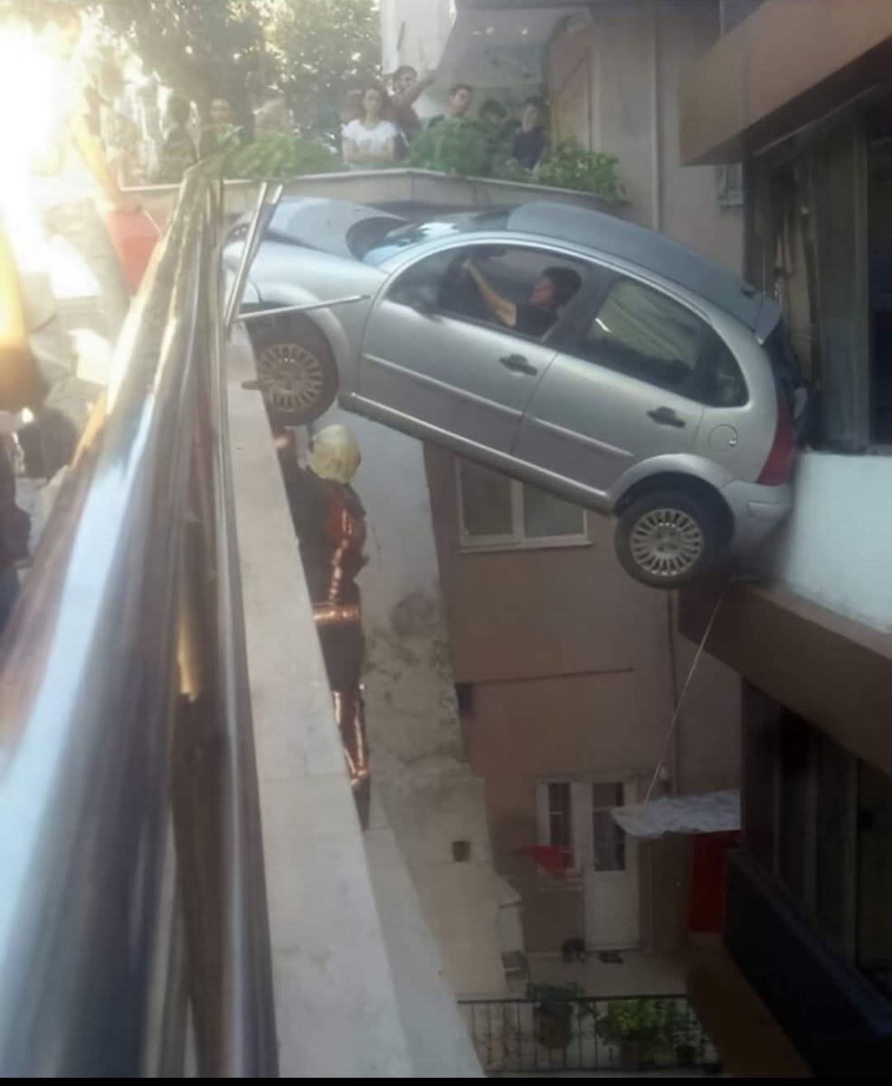 Auto visí na okraji budovy, diváci sledujú incident, záchranári zasahujú pri neobvyklej dopravnej nehode.