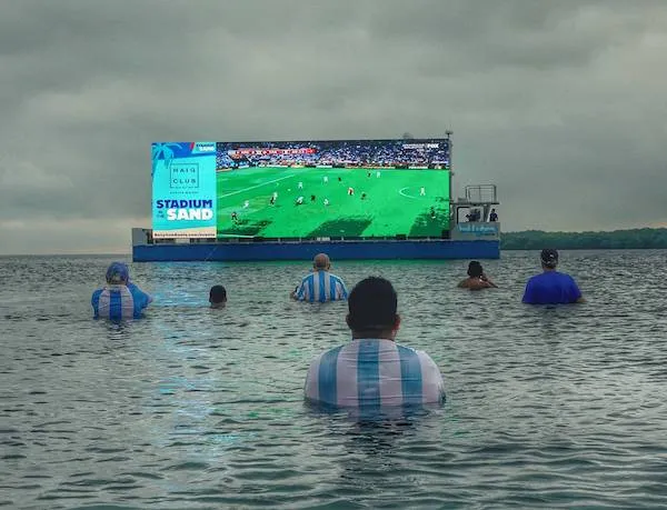 Fanúšikovia sledujú futbalový zápas na veľkej obrazovke uprostred vody, oblečení v modro-bielych dresoch.