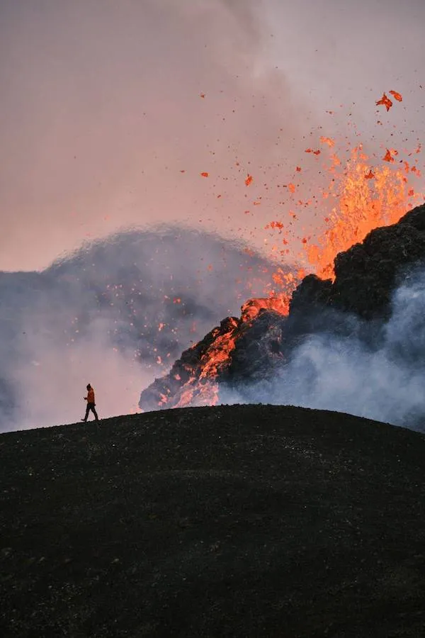 Osoba kráča blízko aktívnej sopky s lávou a dymom v pozadí, dramatická scenéria prírody.