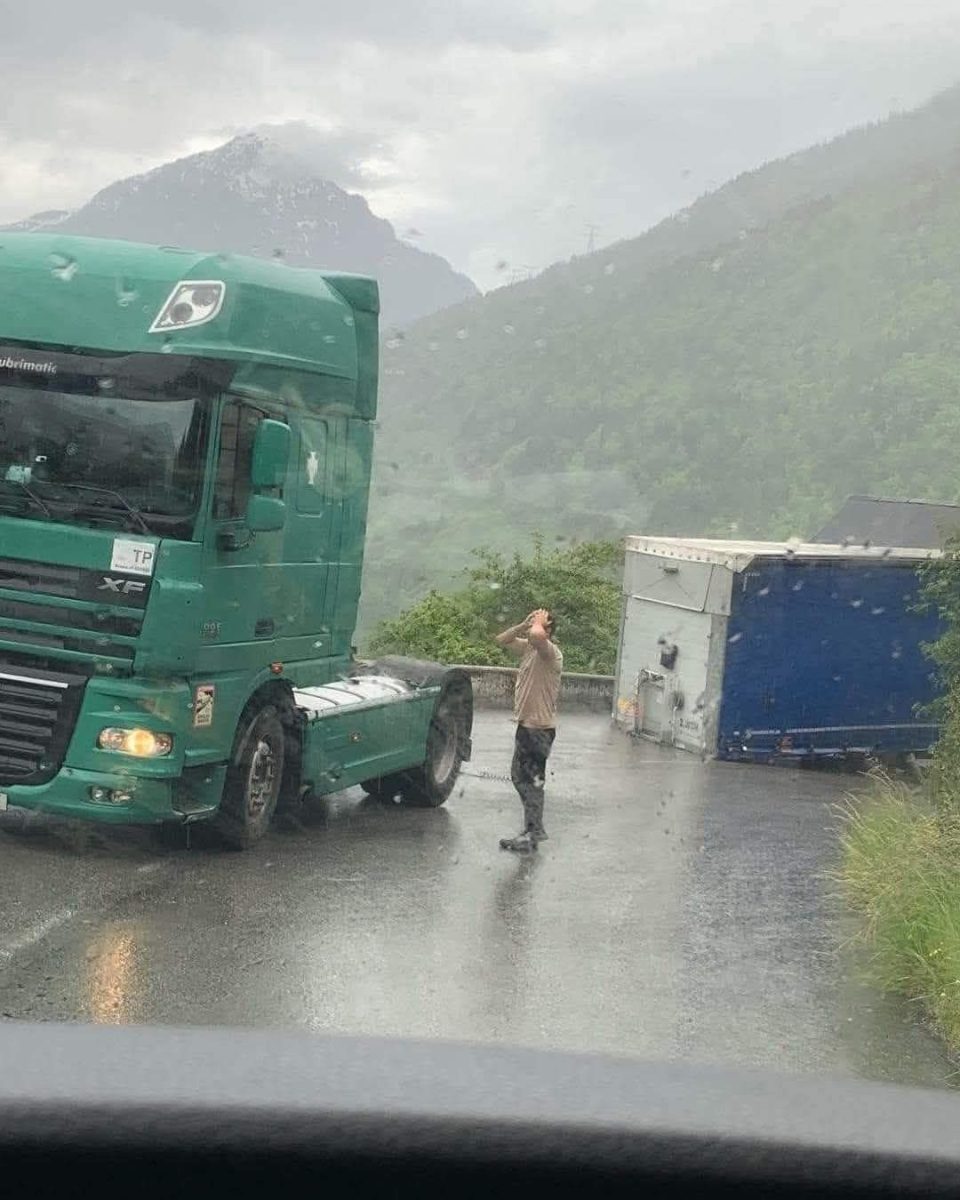 Muž stojí na mokrej ceste pred zeleným kamiónom vedľa prevráteného prívesu, hory v pozadí.