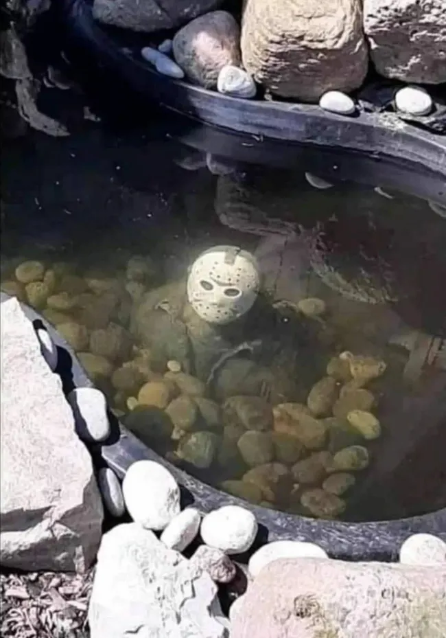 Maska na dne jazierka obklopená kameňmi, evokujúca záhadnú atmosféru v záhradnej dekorácii.