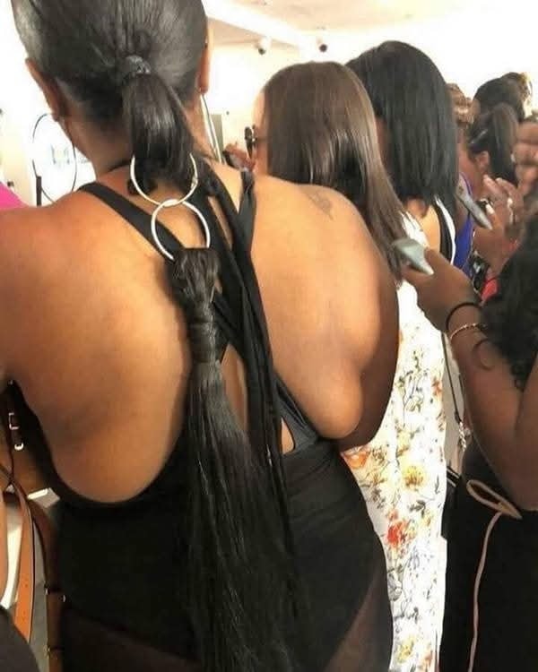 Žena s dlhou vrkočou zviazanou kruhmi stojí v rade v rušnom prostredí, ľudia okolo používajú mobilné telefóny.