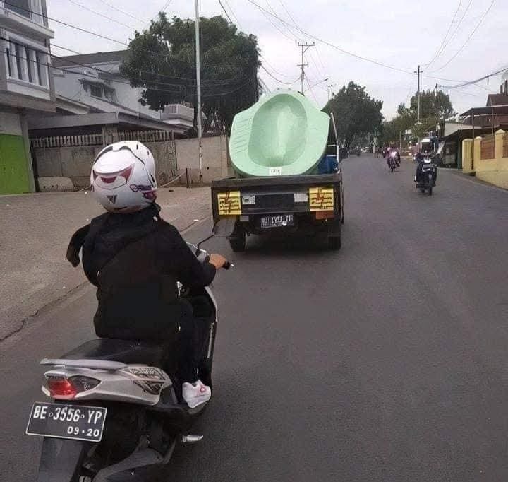 Motorkár sleduje nákladné auto viezúce veľký zelený predmet na ceste v meste.