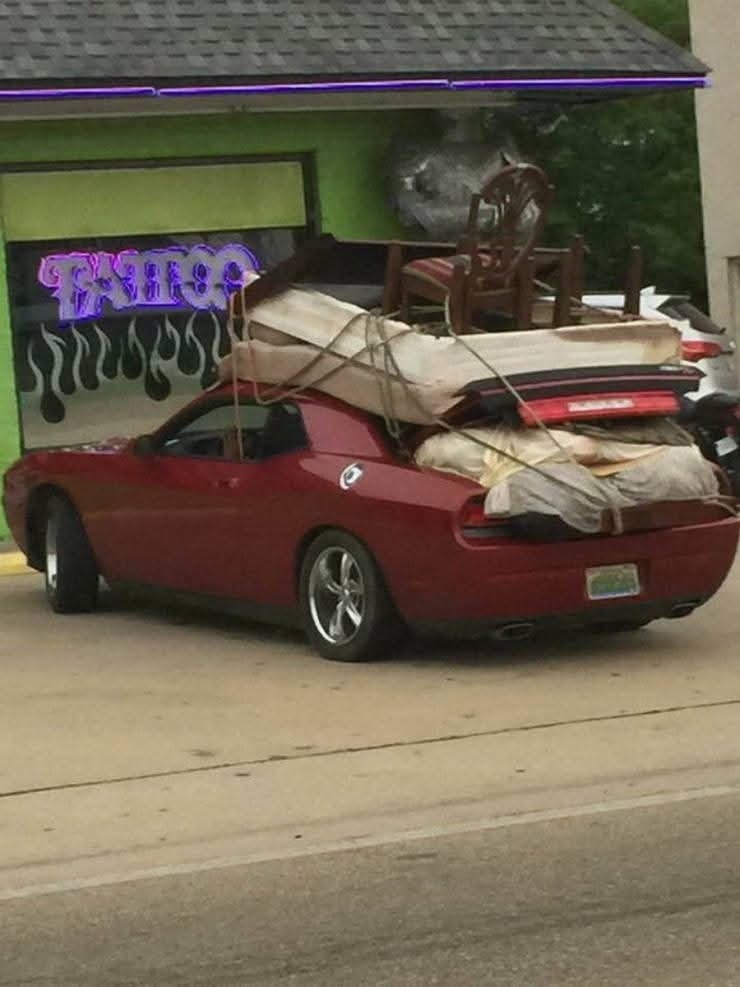 Červené auto naložené nábytkom, matracom a stoličkami upevnenými lanami na streche, zaparkované pred obchodom.