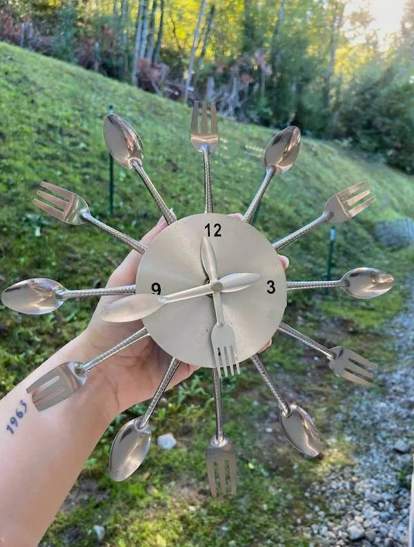 Hodiny z vidličiek a lyžíc držiace v ruke, umiestnené na prírodnom pozadí.