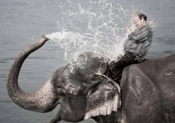 Muž sediaci na slonovi si užíva osviežujúcu sprchu z jeho chobota pri vode.