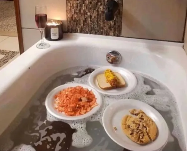 Jedlo na tanieroch plávajúce vo vani plnej peny, vedľa pohár vína a sviečka na okraji vane.