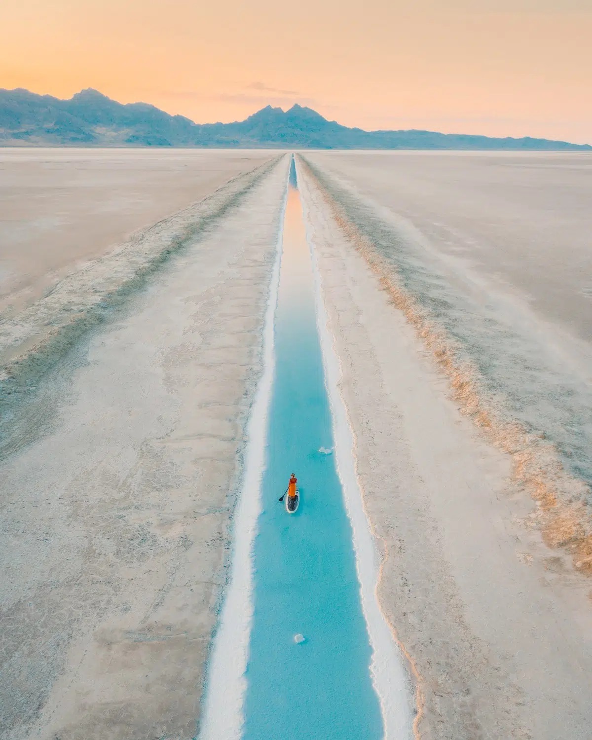 Osoba na paddleboarde na tyrkysovom kanáli uprostred soľnej pláne s horami v pozadí pri západe slnka.