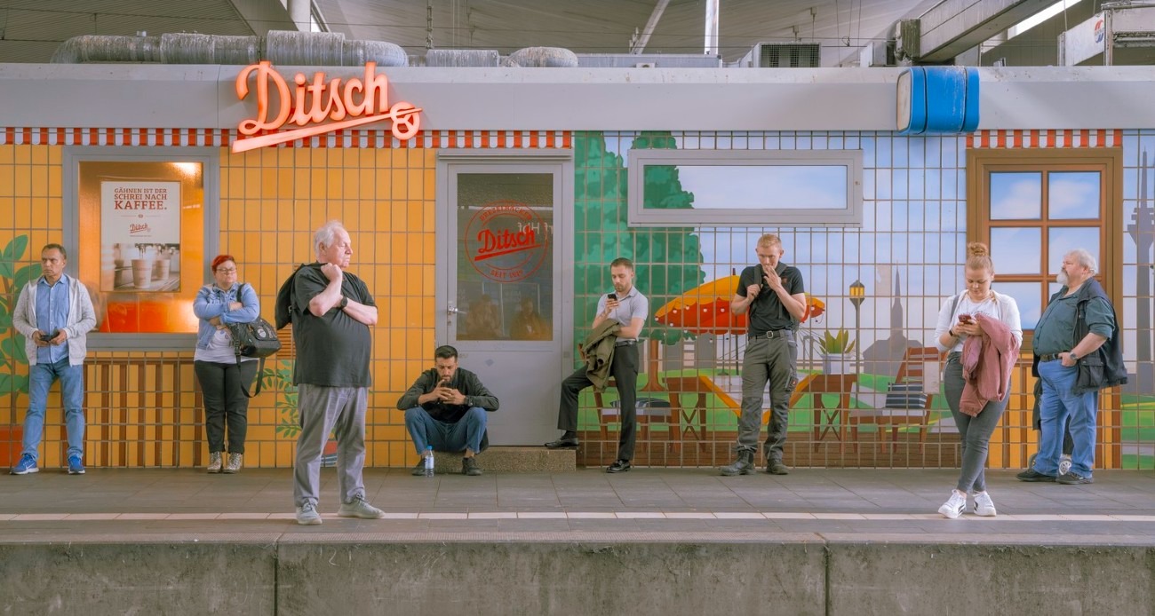 Cestujúci čakajú na vlak pred pestrofarebnou maľbou v stanici pri stánku s nápisom Ditsch.