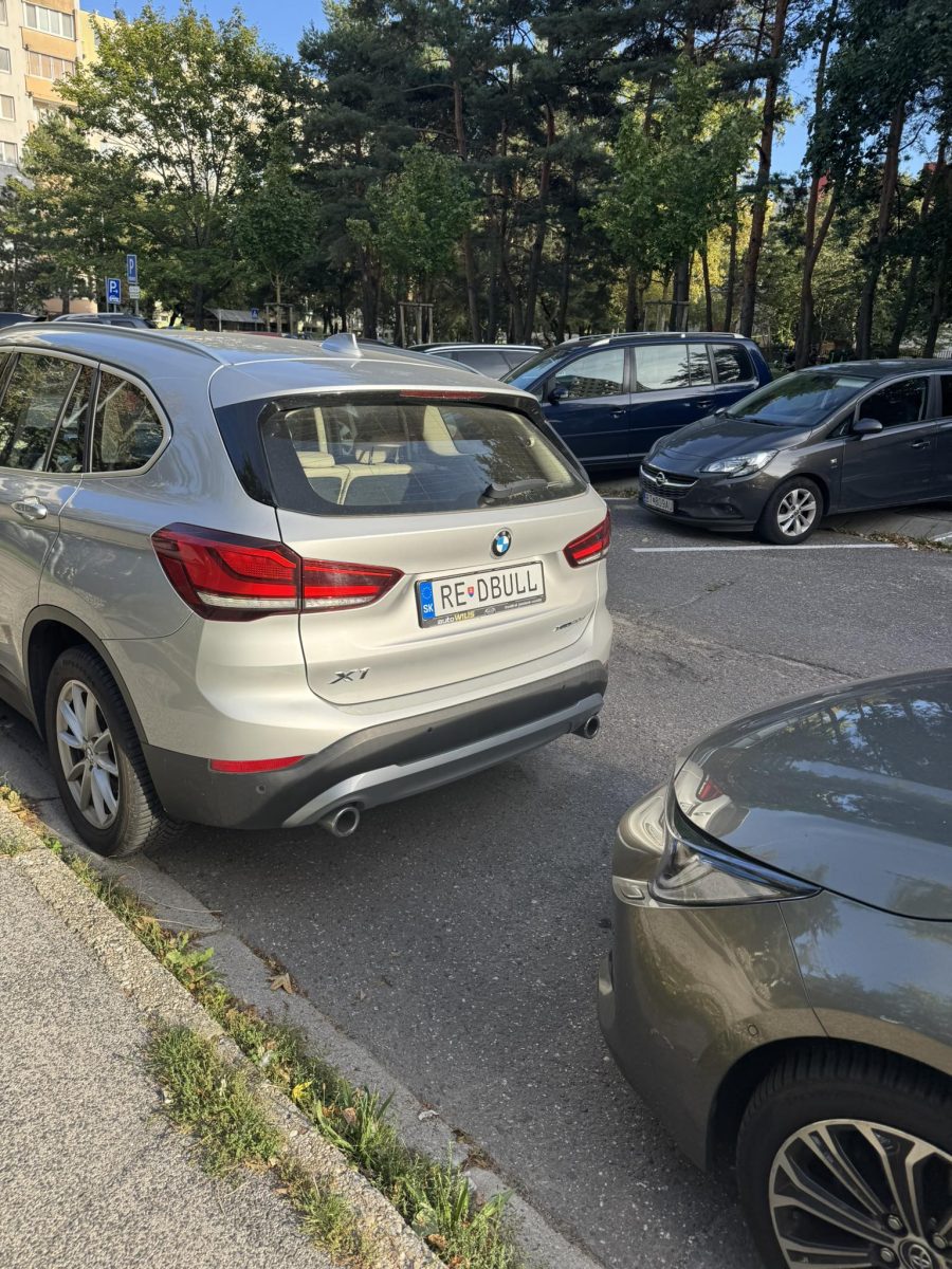 BMW X1 s vtipnou poznávacou značkou parkuje na parkovisku obklopenom stromami.