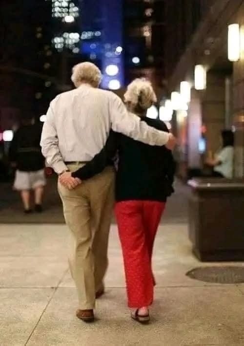 Starší pár sa prechádza večerným mestom, objímajúc sa a užívajúc si spoločné chvíle.