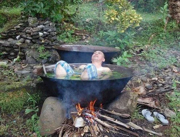 Muž relaxuje v horúcej vode v prírode, leží v kovovej vani nad ohňom, obklopený zeleňou.