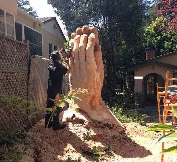 Umelec pracuje s motorovou pílou na veľkej drevenej soche zložených rúk v záhrade.