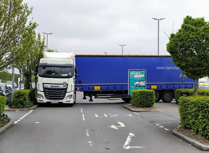 Kamión blokujúci cestu na parkovisku medzi zelenými kríkmi a lampami.