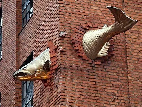 Zlatá ryba prechádzajúca tehlovou stenou budovy, umelecká inštalácia na tehlovej fasáde.